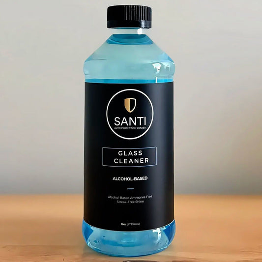 A bottle of Santi Alcohol-Based Glass Cleaner with a black label and black cap, filled with a bright blue liquid, sitting on a wooden surface against a neutral gray background.