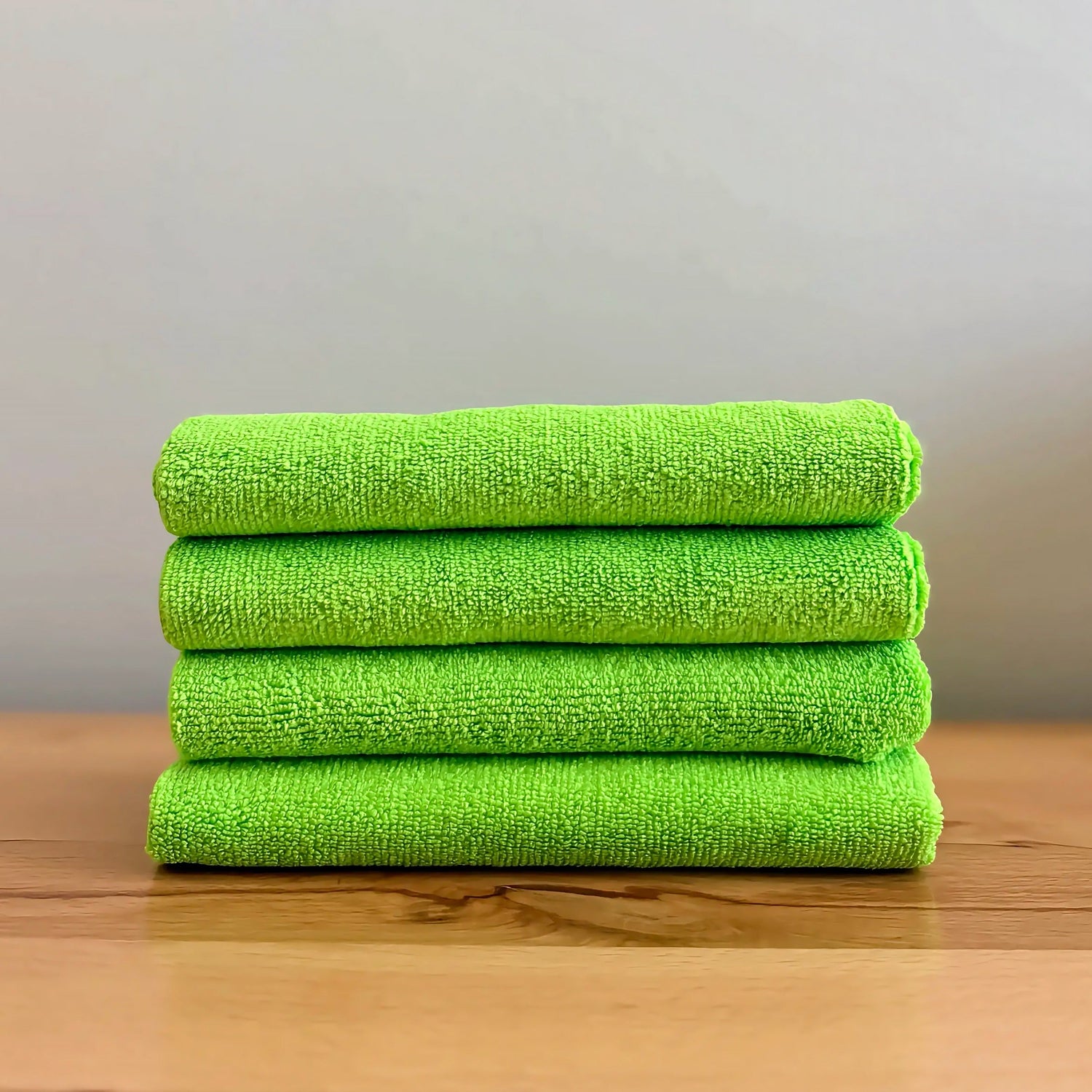 Four bright green microfiber cleaning towels stacked neatly on a wooden tabletop against a neutral gray background.