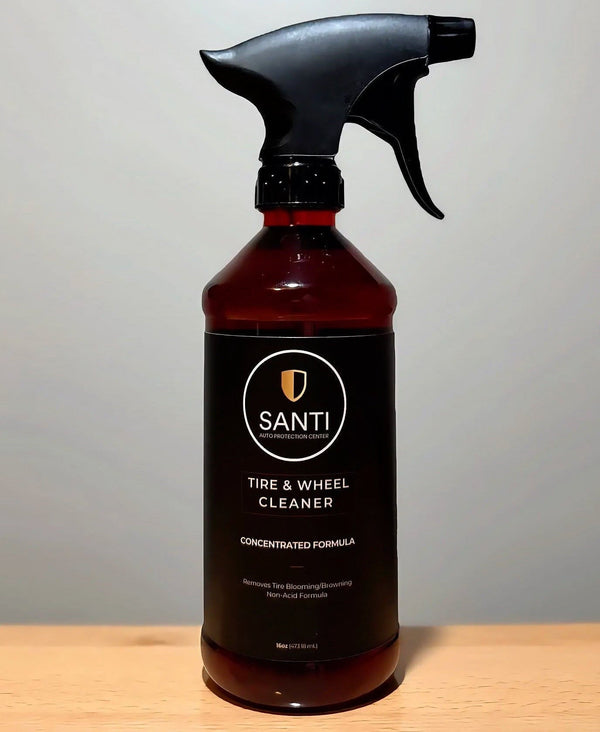 A bottle of Santi Tire & Wheel Cleaner with a black spray trigger and dark amber liquid inside, standing on a light wood surface against a neutral gray background.
