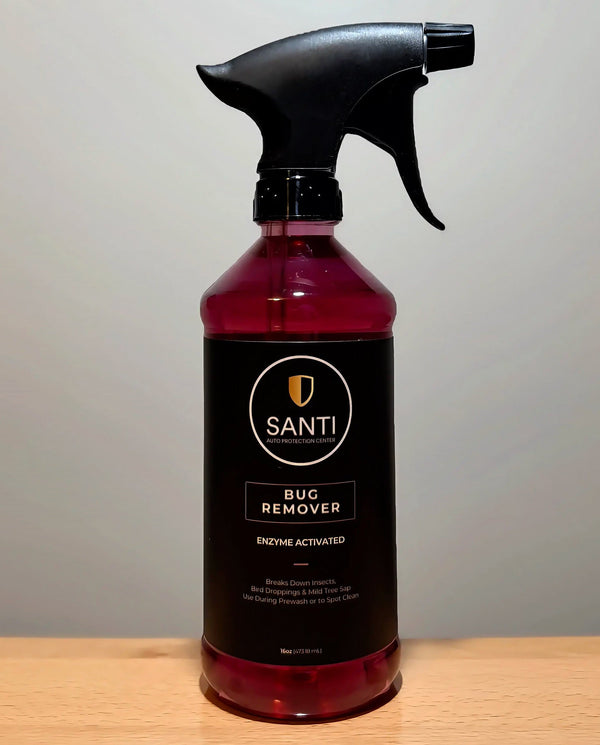 A bottle of Santi Bug Remover insecticide with a black spray trigger top and a dark label, filled with a deep red liquid, sitting on a light wood surface against a neutral gray background.
