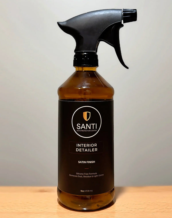 A bottle of Santi Interior Detailer spray cleaner with a black trigger sprayer and amber glass bottle, sitting on a light wood surface against a neutral gray background.