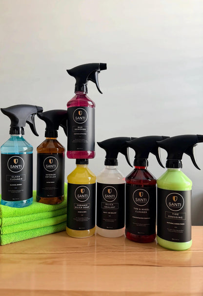 A set of SANTI brand glass and window cleaning spray bottles in various colors, arranged on a wooden table with a stack of green microfiber cloths. The bottles feature black spray triggers and black labels with white text.