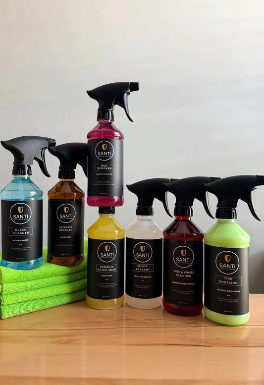 A set of SANTI brand glass and window cleaning spray bottles in various colors, arranged on a wooden table with a stack of green microfiber cloths. The bottles feature black spray triggers and black labels with white text.