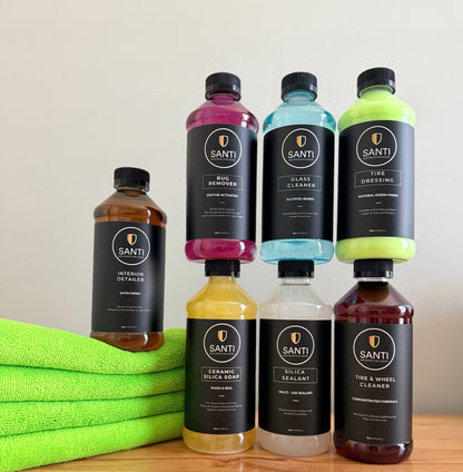 Six bottles of SANTI car care products arranged on a wooden surface against a light wall, with a bright green towel on the left. The bottles contain amber, pink, blue, yellow, white, and dark red liquids and have black labels with gold SANTI branding.