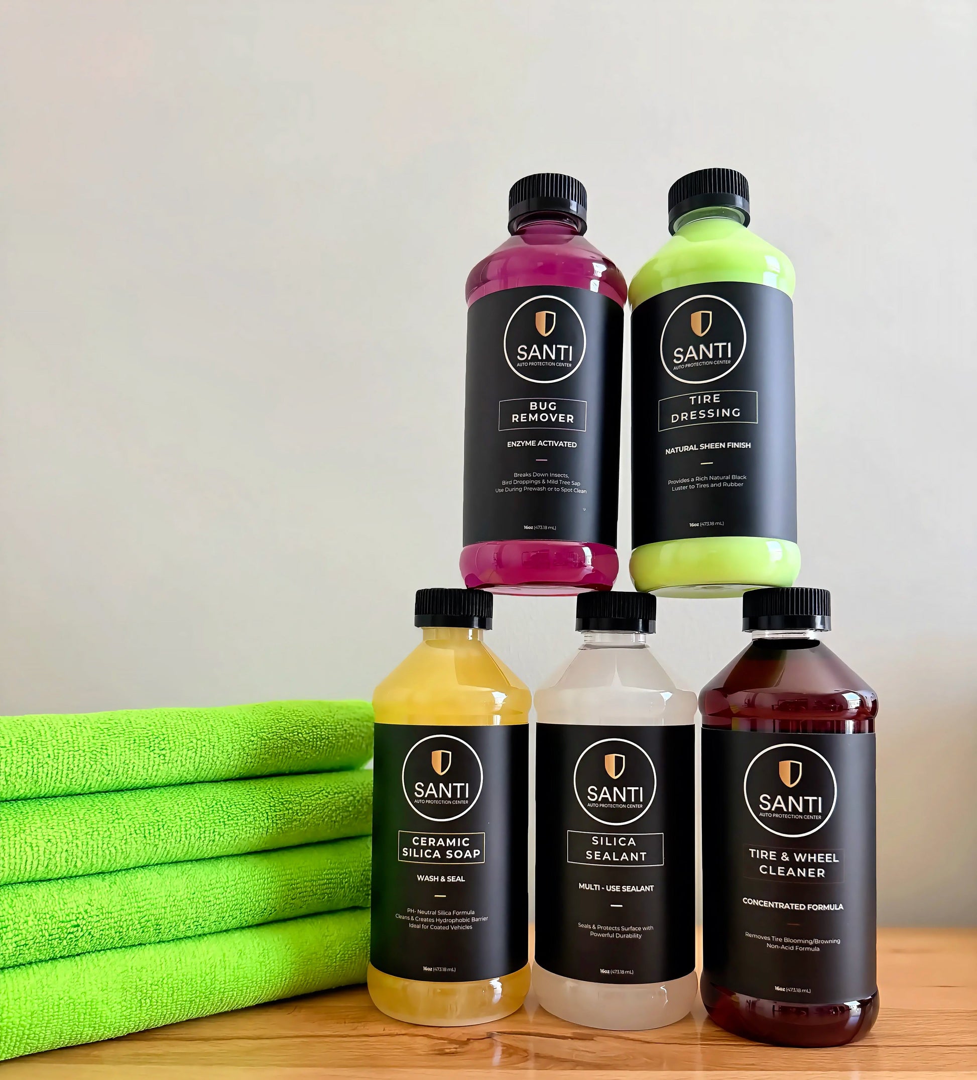 Four Santi branded car care bottles arranged in a pyramid on a wooden surface, with folded bright green towels on the left. The bottles contain pink, neon green, yellow, and dark reddish-brown liquids and have black labels with white text.