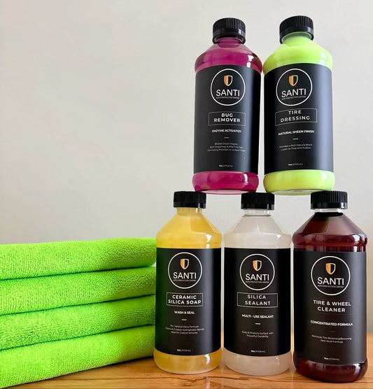 Four Santi branded car care bottles arranged in a pyramid on a wooden surface, with folded bright green towels on the left. The bottles contain pink, neon green, yellow, and dark reddish-brown liquids and have black labels with white text.