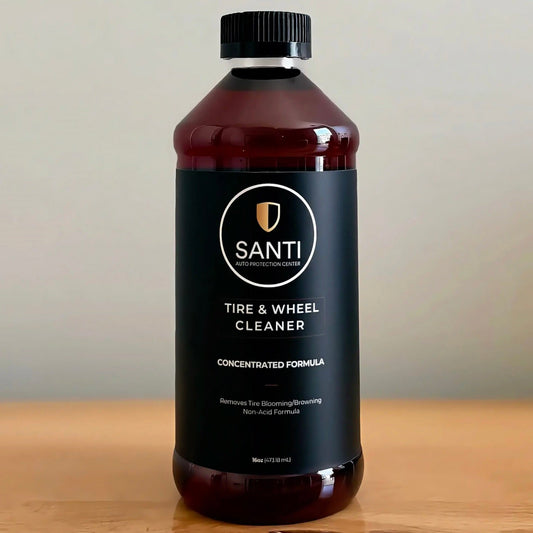 A bottle of Santi Tire & Wheel Cleaner with a black cap and black label, placed on a wooden surface against a neutral beige background.