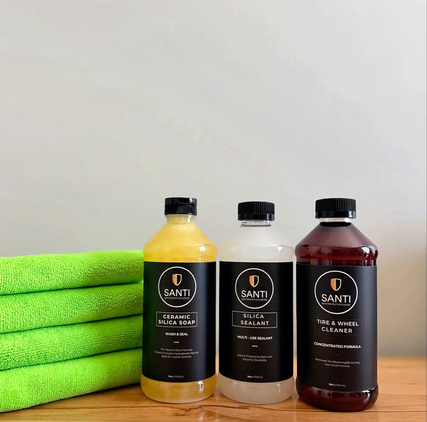 Three bottles of Santi car care products lined up on a wooden surface next to a stack of bright green microfiber towels. The bottles contain yellow, white, and dark reddish-brown liquids and are labeled Ceramic Silicon Soap, Silica Sealant, and Tire & Wheel Cleaner.