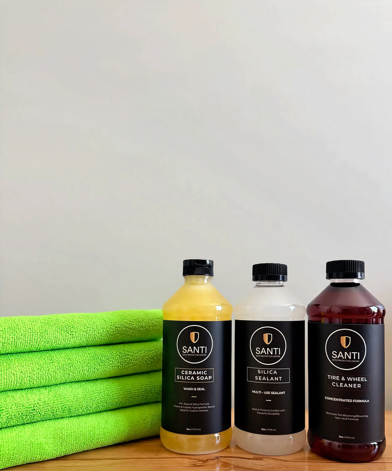 Three bottles of Santi car care products lined up on a wooden surface next to a stack of bright green microfiber towels. The bottles contain yellow, white, and dark reddish-brown liquids and are labeled Ceramic Silicon Soap, Silica Sealant, and Tire & Wheel Cleaner.