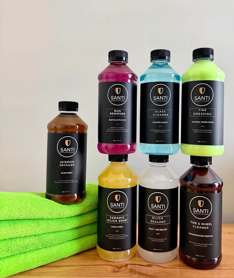 Six bottles of SANTI car care products arranged on a wooden surface with a bright green towel. The set includes Interior Detaler, White Remover, Glass Cleaner, Tire Dressing, Ceramic Silica Soap, Silica Sealant, and Tire & Wheel Cleaner in various translucent colors.