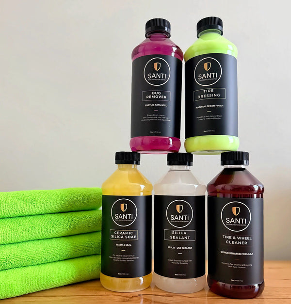Four bottles of SANTI car care products arranged on a wooden surface with a stack of green microfiber towels on the left. The bottles contain pink, neon green, yellow, and dark red liquids and have black labels with the SANTI brand name.