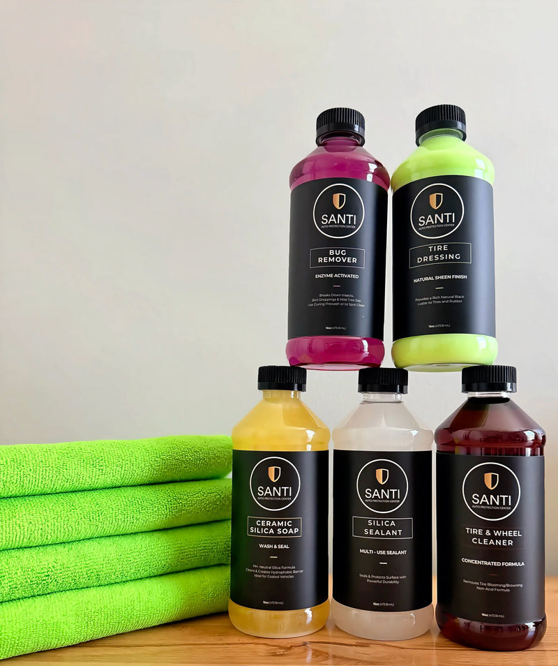 Four bottles of SANTI car care products arranged on a wooden surface with a stack of green microfiber towels on the left. The bottles contain pink, neon green, yellow, and dark red liquids and have black labels with the SANTI brand name.
