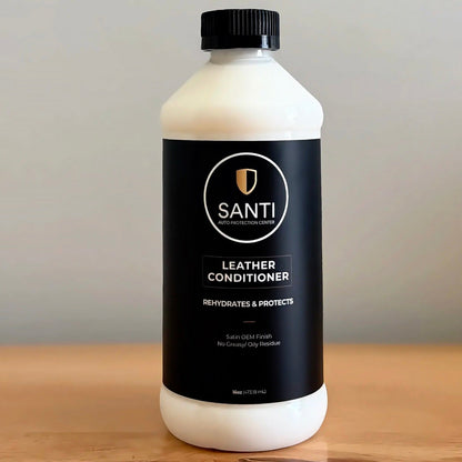 A bottle of Santi Leather Conditioner with a black label and black cap, sitting on a wooden surface against a neutral gray background.