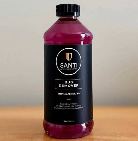 A bottle of Santi Bug Remover with a black label and black cap, filled with a deep magenta liquid, set on a wooden surface against a neutral beige background.