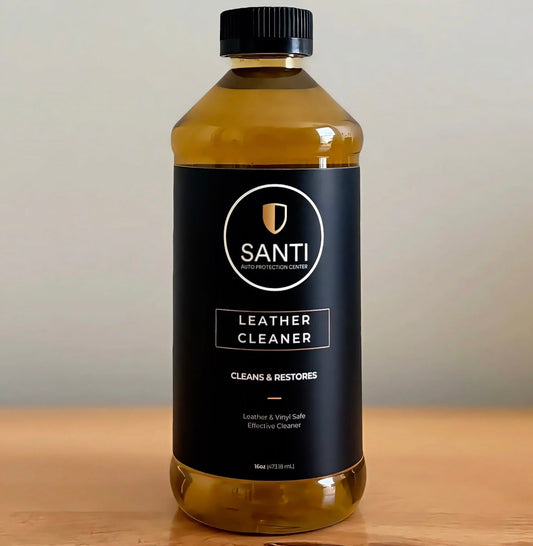 A bottle of Santi Leather Cleaner with a black label and black cap, filled with amber-colored liquid, sitting on a wooden surface against a neutral gray background.