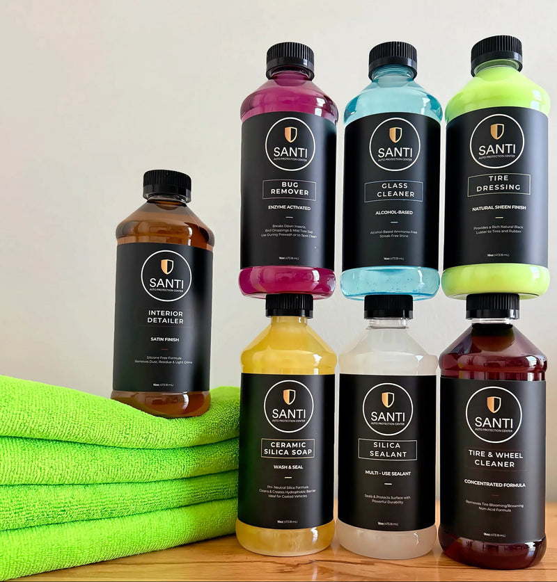 Eight Santi brand liquid car care products in clear bottles with black caps and black labels, arranged on a wooden surface with a bright green towel. The products include interior detailing, tire dressing, glass cleaner, brake cleaner, ceramic silver soap, silica sealant, and the a wheel cleaner.