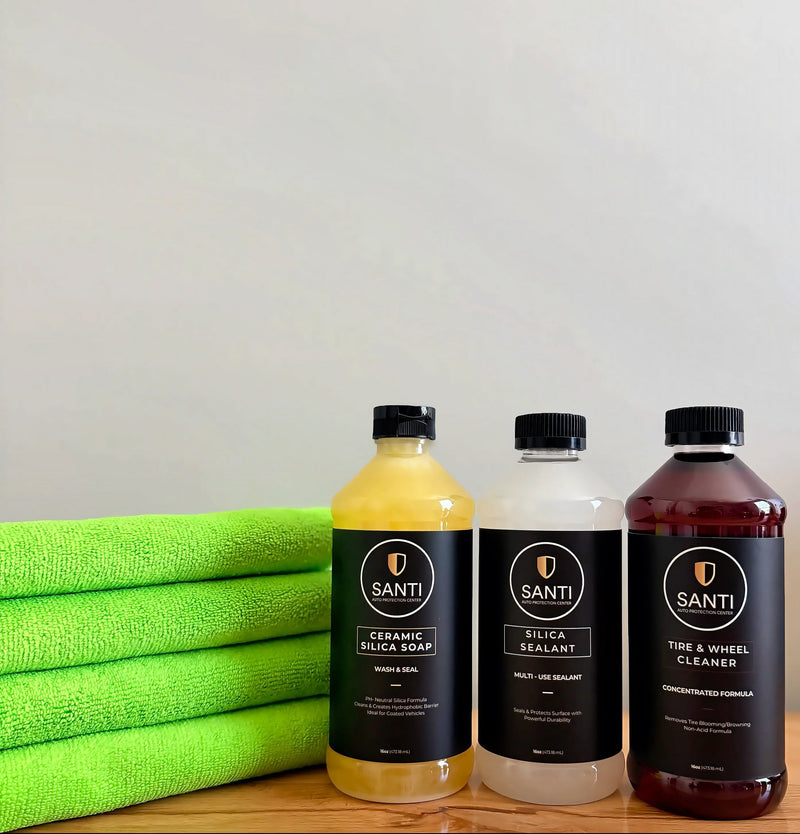 Three bottles of Santi car care products lined up on a wooden surface next to a stack of bright green microfiber towels. The bottles contain yellow, white, and dark reddish-brown liquids and are labeled Ceramic Silicon Soap, Silica Sealant, and Tire & Wheel Cleaner.